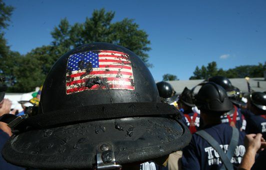 Firefighter helmet with worn American flag sticker, blue sky, trees, and other firefighters in the background.