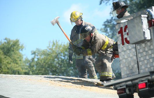 Firefighters on a roof with an ax; smoke visible, under a bright blue sky.