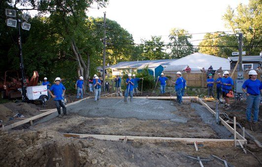 Construction workers in blue shirts and white helmets pouring concrete for a foundation. Outdoor setting.