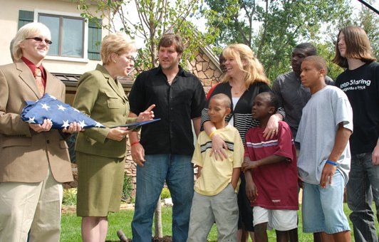 Group outdoors: woman presenting folded flag to family in front of a house.