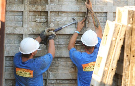 Two construction workers in blue shirts and white hard hats removing a brick-patterned form with hammers.