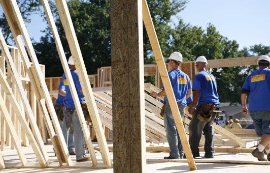 Construction workers in blue shirts and hard hats building a wood-framed structure outdoors.