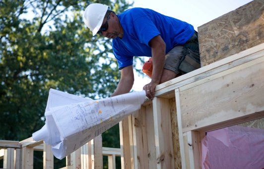 Construction worker on framing, examining blueprints. He wears a hard hat and blue shirt. Outdoors.