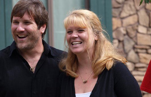 Man and woman smiling, outdoors. The man wears a black shirt, the woman has blonde hair and a dark sweater.
