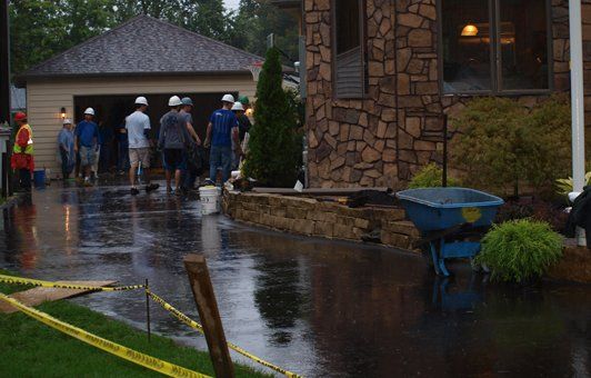 Workers in hard hats on a wet driveway, near a stone building and a garage, with a wheelbarrow.