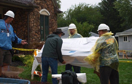 Four people in hard hats and rain gear lifting a large, white object outside a house.