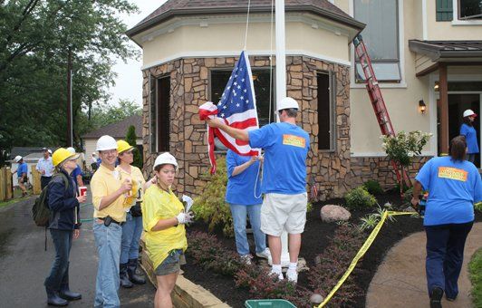 People raising an American flag on a flagpole outside a building. Several wear hard hats, sunny day.