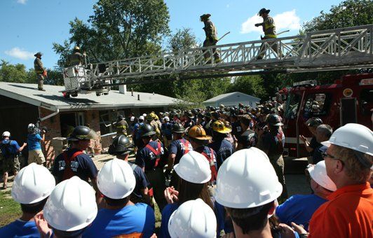 Firefighters on roof and ladder, smoke visible, with crowd of people in hard hats looking on.