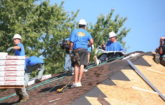 Roofers in blue shirts and hard hats install shingles on a rooftop on a sunny day.