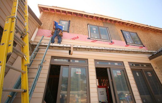 Construction worker installing siding on a two-story building. A ladder provides access.