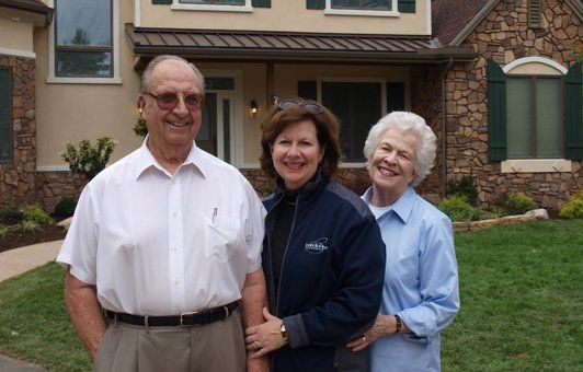 Three people smiling in front of a house with stone accents, one person is wearing a white shirt and glasses.