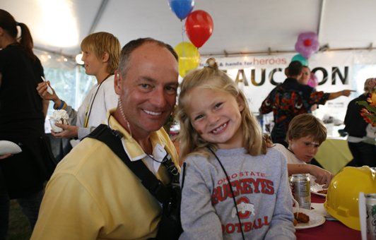 Man and young girl smiling at a table, both looking at the camera. Balloons, other people in background.
