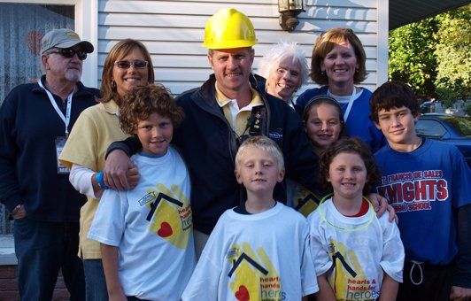 Group of people, including children, pose in front of a house, some wearing construction-related items.
