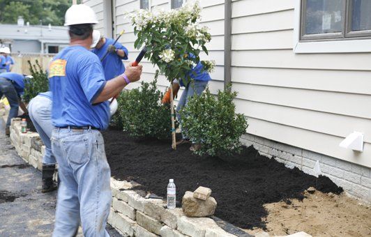 Landscapers mulching a garden bed along a building's side, using tools and materials.