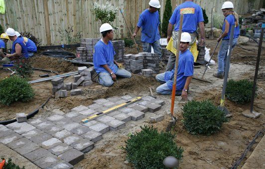 Construction workers laying paving stones in a garden, wearing blue shirts and hard hats.