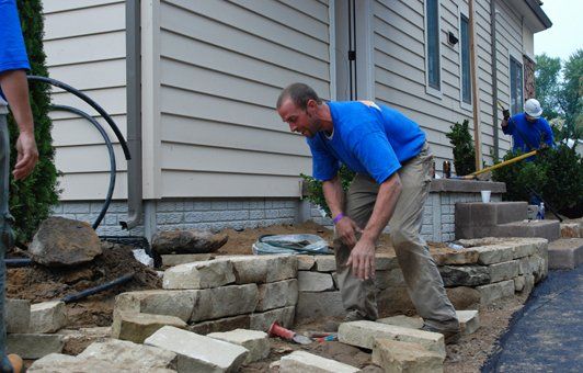 Man in blue shirt building a stone wall near a house. Another worker in background.