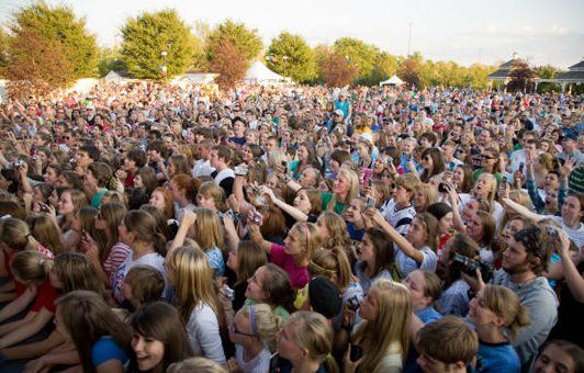 Large crowd outdoors, many people with arms raised, likely at a concert or event, trees in background.