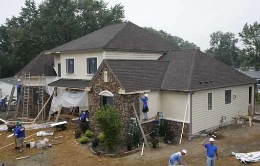 New house under construction, workers around exterior. Beige siding, stone facade, brown roof.