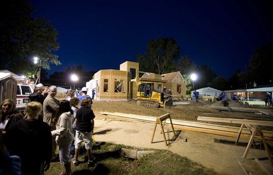 People watching a home being built at night, construction workers, and a yellow excavator.