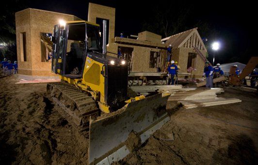 Yellow bulldozer moving a wooden house at night, construction workers nearby.