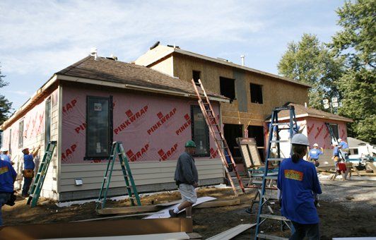 Construction workers building a two-story house, applying pink wall wrap. Ladders and materials are present.