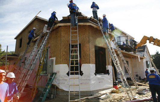 Construction workers on a house roof, using ladders. Building exterior is wood-framed.