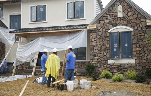 Construction workers working on a house exterior. White tarps cover a porch, stone and stucco siding visible.
