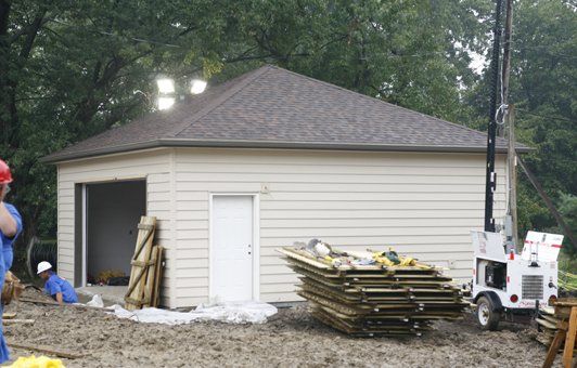 Garage under construction with tan siding, brown roof, white door, and workers present.