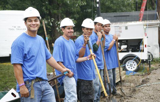 Six people in blue shirts and hard hats, holding shovels. Outdoors, construction site.