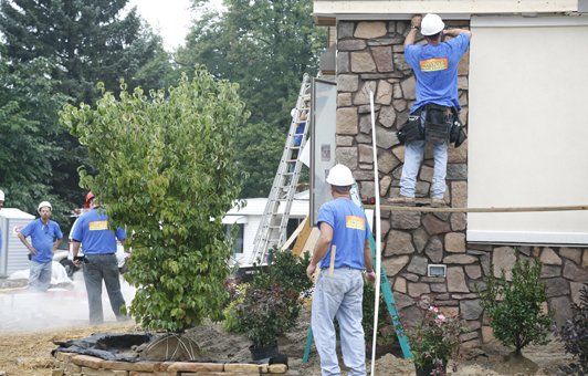 Construction workers installing stone siding on a building exterior. Several wear blue shirts and hard hats.