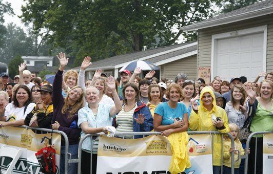 Crowd of people waving behind a barrier. Some have umbrellas. Houses and a garage are in the background.