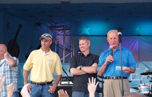 Four men on stage: one with a microphone, others standing. Blue backdrop, crowd in foreground.