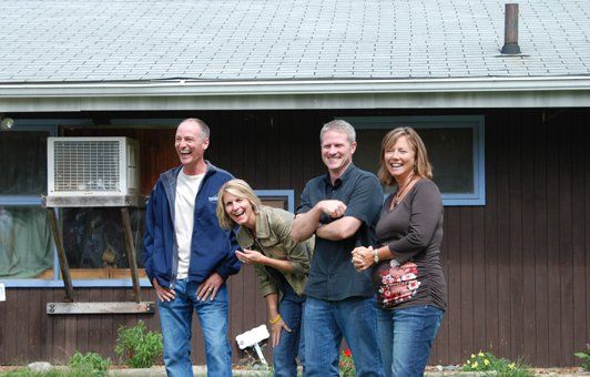 Four smiling people standing in front of a brown house. Two have arms crossed. Overcast sky.