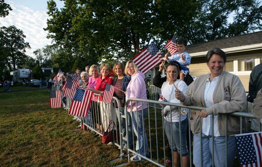 People line up behind a metal fence, holding American flags. Some are smiling, in an outdoor setting.