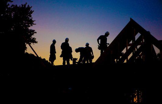 Silhouetted construction workers on rooftop at dusk.