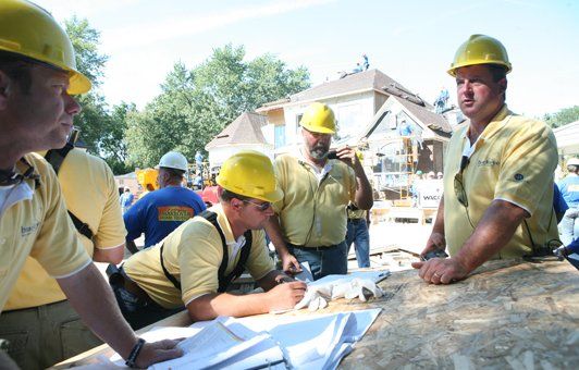 Construction workers in yellow hard hats and shirts reviewing blueprints outside a house.