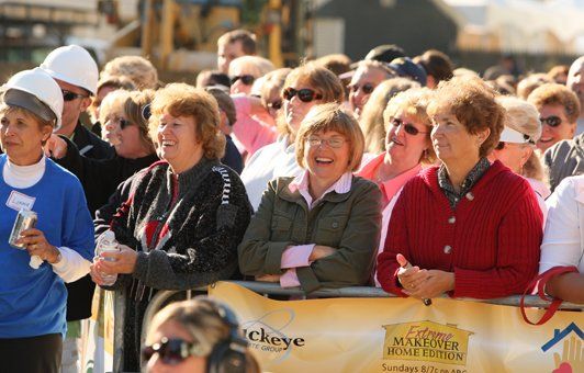 A crowd of people watches an outdoor event, some smiling and clapping. Many wear sunglasses and casual clothing.