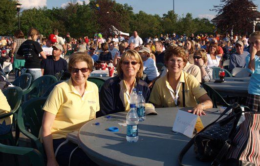 Three women at a table smiling at a crowd at an outdoor event.