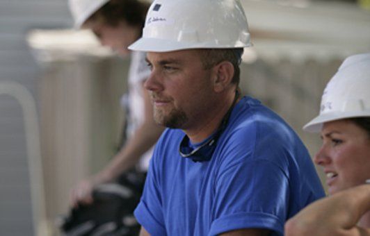 Man in blue shirt and hard hat, looking off-screen. Others wear hard hats, in a construction setting.