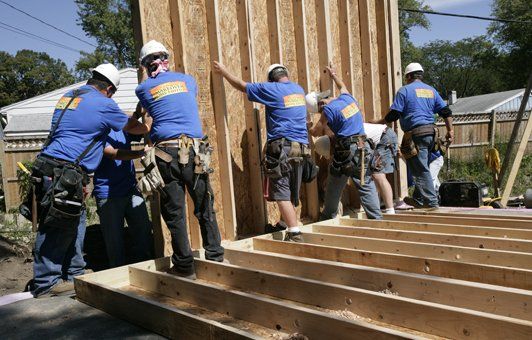 Construction workers lift a wall panel onto a wooden frame at a building site.