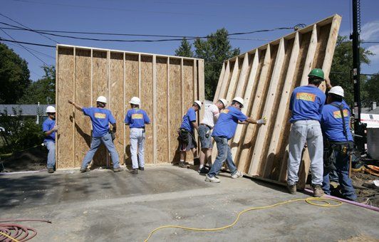 Construction workers in blue shirts and white helmets lifting a wall panel outdoors.