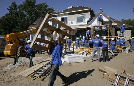 Construction workers in blue shirts and hard hats building a two-story house.
