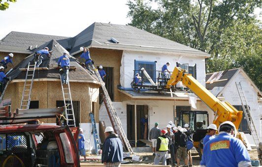 Construction workers on a two-story house, installing siding and roofing. Yellow lift, ladders, and red truck visible.