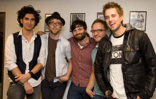 Five men posing closely together in front of a wall with framed art. They wear casual clothing and smile.