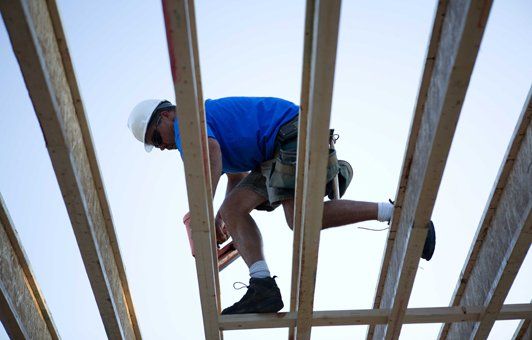 Construction worker on roof beams, reaching, blue shirt, white helmet, sunny sky.
