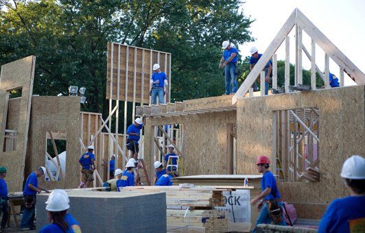 Construction workers in blue shirts and hard hats building a wood-framed house outdoors.