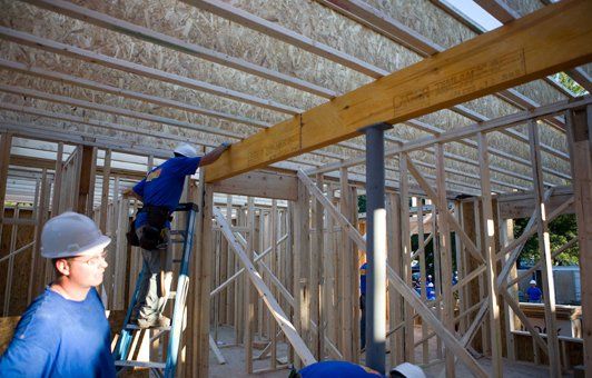 Construction workers building a wooden structure. One on a ladder, installing a beam. Interior framing visible.