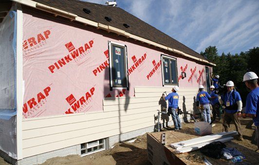 Construction workers installing siding on a house, pink wrap visible, sunny outdoor setting.