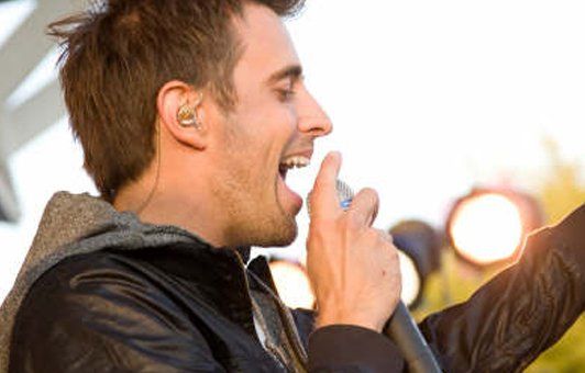 Man singing into a microphone, outdoors, wearing a black jacket, open-mouthed with excitement.