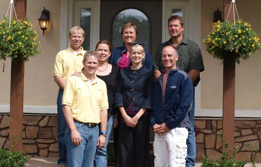 Group of eight people posing on the steps of a house with hanging baskets; some smiling, some looking at camera.